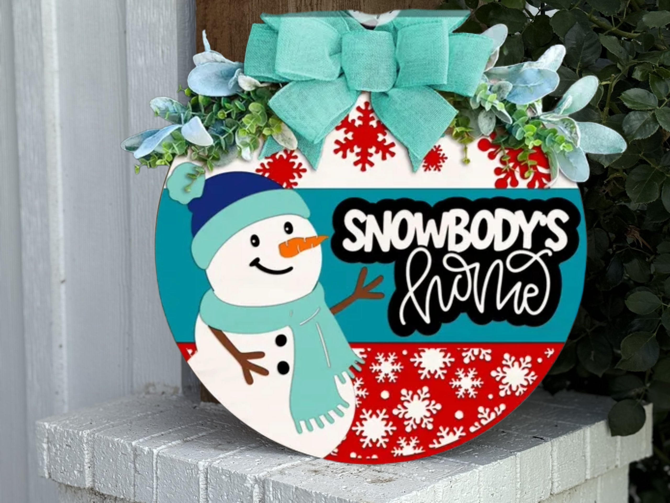 A round winter-themed sign with a snowman, red snowflakes, blue stripes, the words “Snowbody’s Home,” and a green bow with greenery on top, displayed on a white brick surface by a leafy plant.