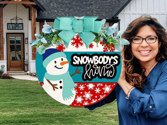 A smiling woman with glasses holds a round winter-themed sign that reads Snowbodys Home, decorated with a snowman, red snowflakes, greenery, and a blue bow, standing in front of a house.