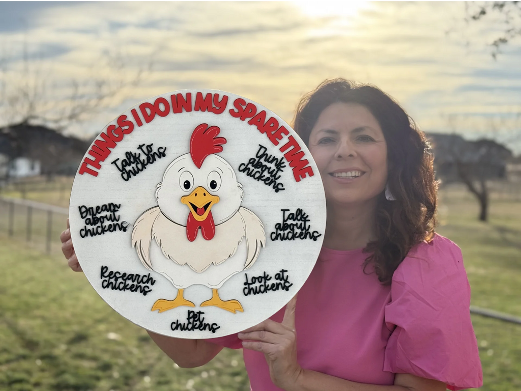 A woman smiles, holding a decorative sign depicting a cartoon chicken surrounded by text. The words read: Things I do in my spare time, followed by Talk about chickens, Dream about chickens, Talk to chickens, Look at chickens, Research chickens, Bet on chickens. She wears a pink shirt.