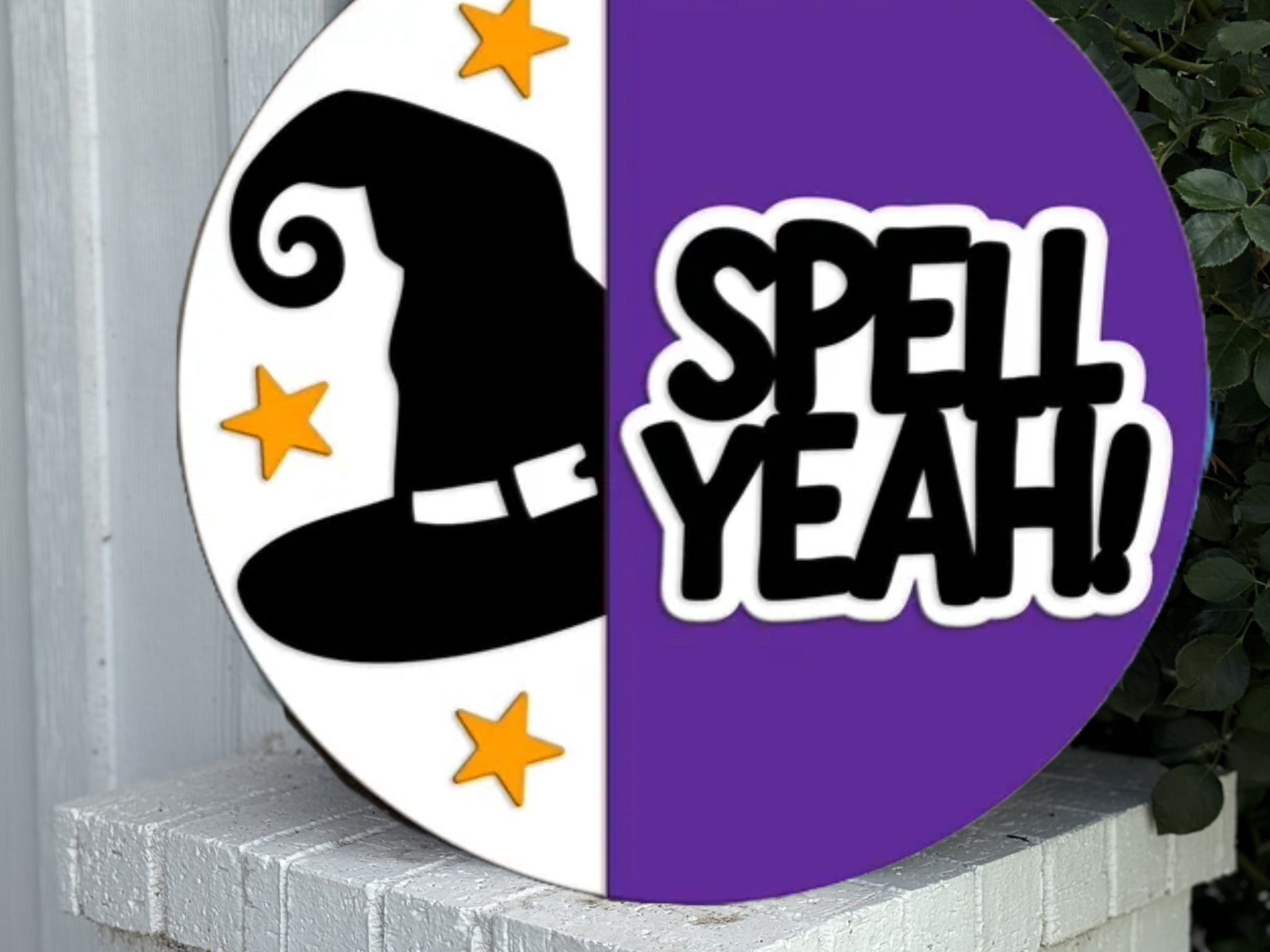 A round sign split in half; the left side is white with a black witch hat and orange stars, the right side is purple with the bold phrase “SPELL YEAH!” in black and white lettering.