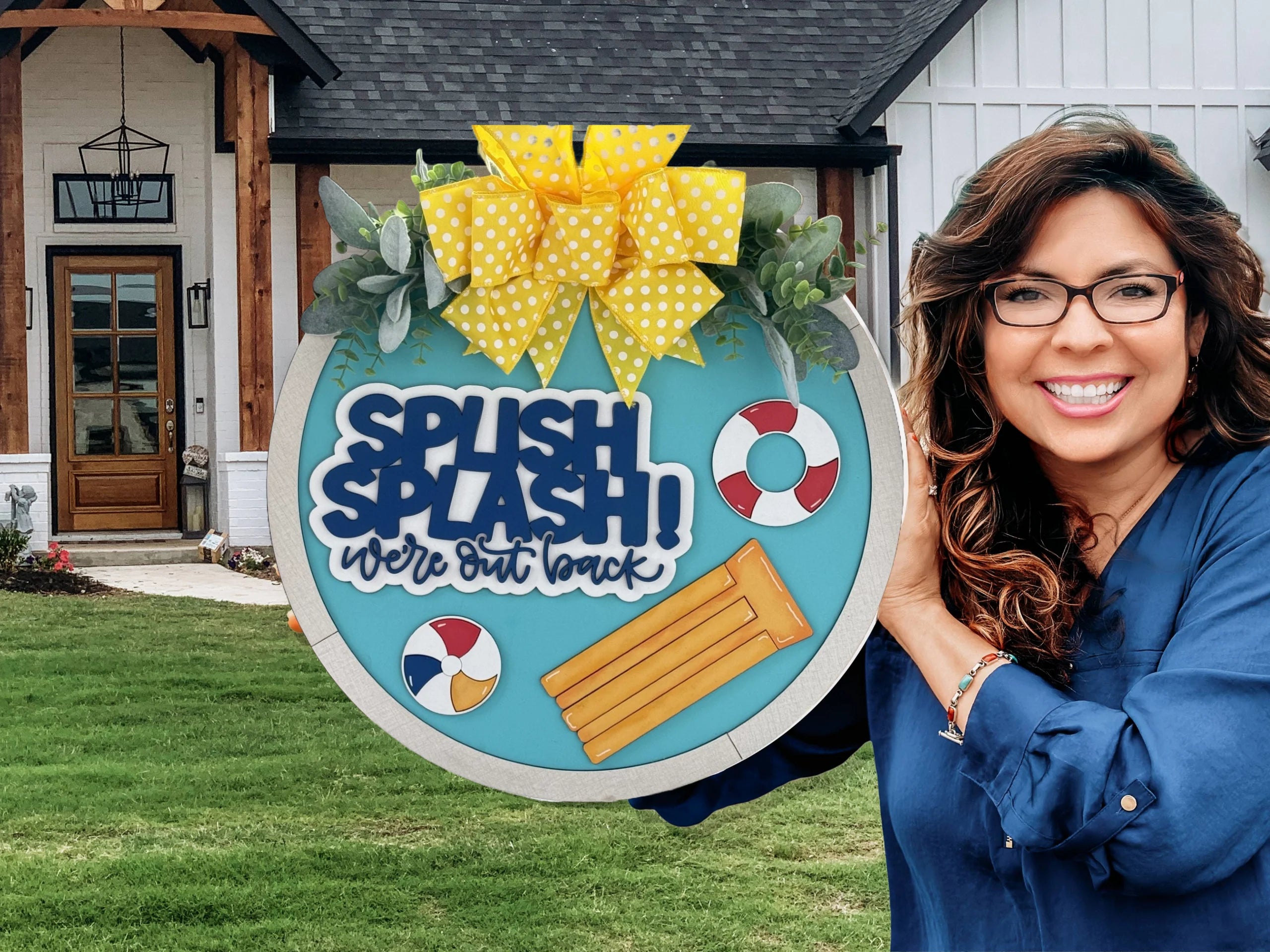 Woman smiling and holding a decorative sign. The sign is circular, blue, with SPLISH SPLASH! were out back in bold letters. It features a pool float, beach balls, and life rings. A yellow polka-dot ribbon and greenery adorn the top. The background shows a house with a wooden porch.