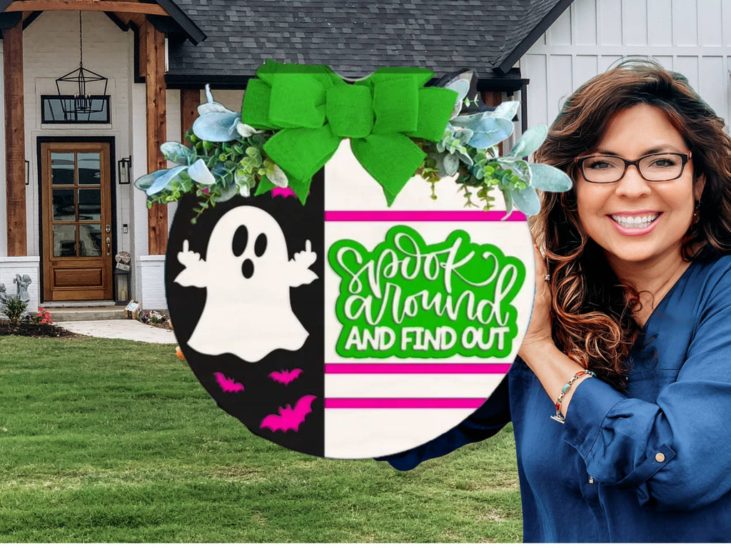 A smiling woman with wavy brown hair and glasses holds a round Halloween sign outside a house. The sign has a ghost, bats, a green bow, and the words Spook around AND FIND OUT in green and white.