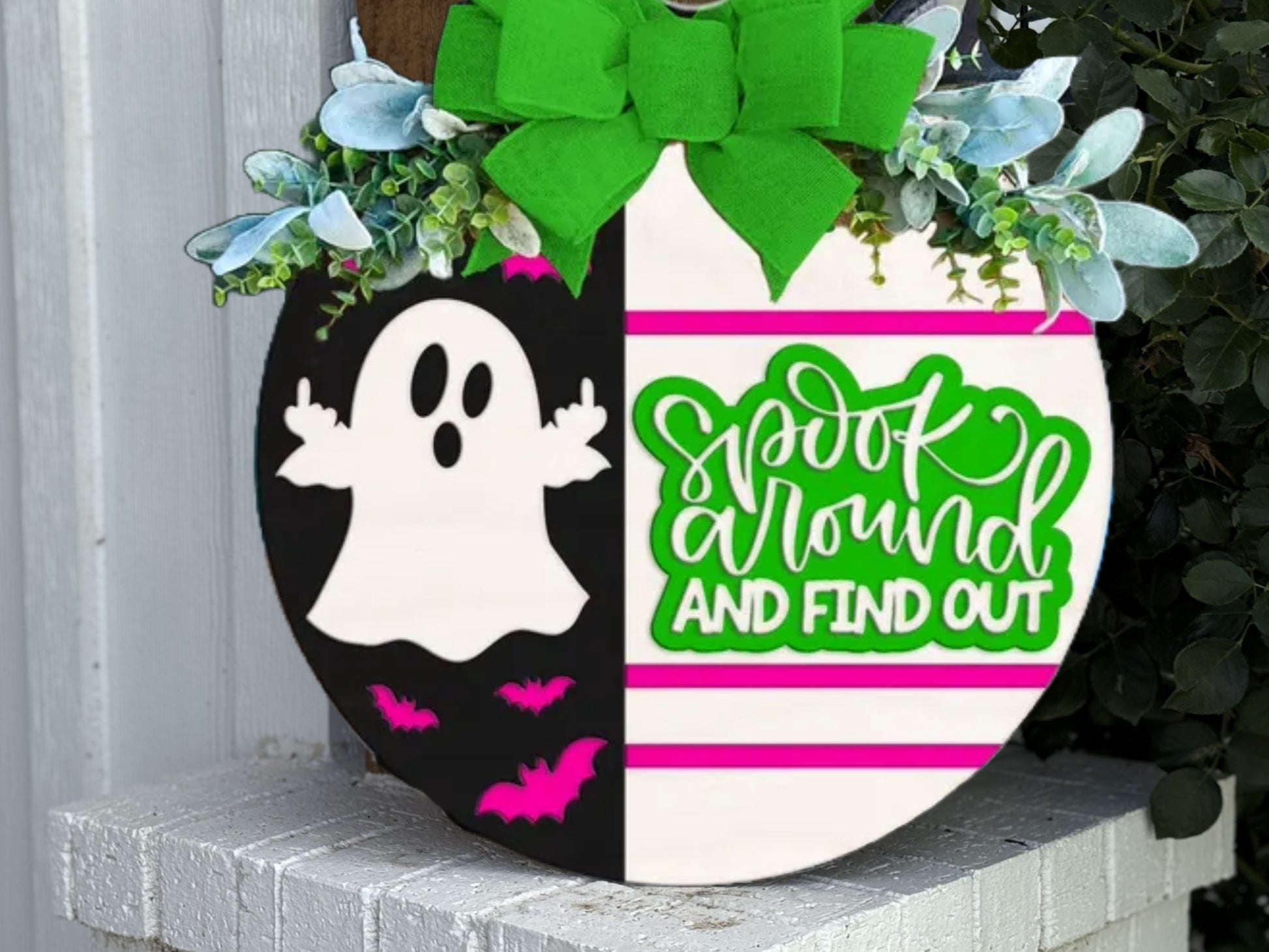 A round sign with a green bow and flowers on top. The left side shows a cartoon ghost and pink bats on a black background; the right side says spook around AND FIND OUT in green and white.