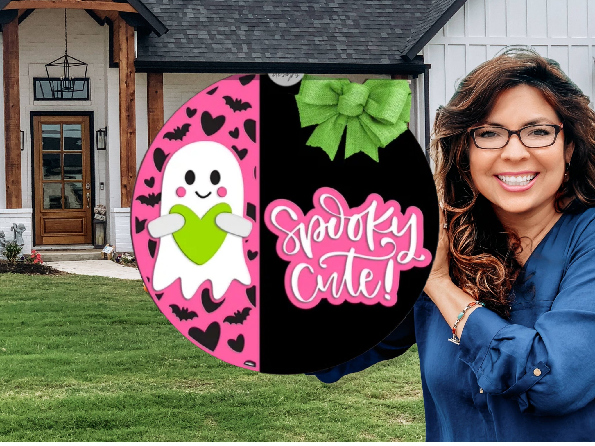 A smiling woman stands in front of a house holding a round sign with a pink ghost holding a green heart on one half and the words Spooky Cute! with a green bow on the other half.