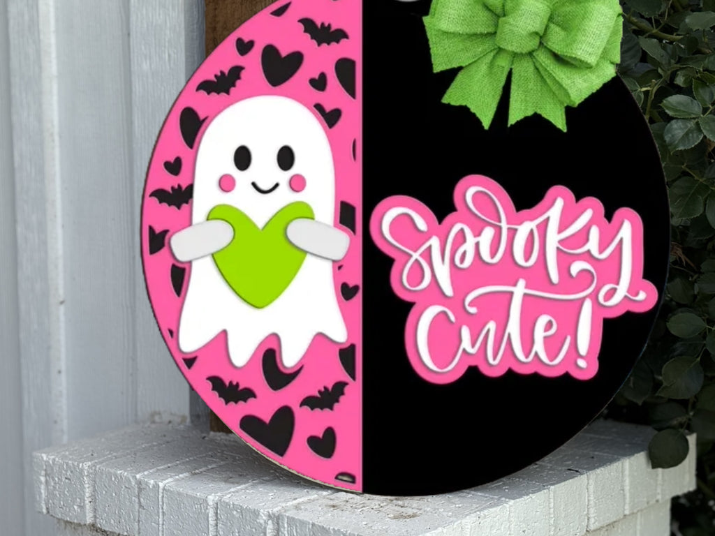 A round sign half pink and half black. The pink side shows a smiling ghost holding a green heart, surrounded by hearts and bats. The black side says Spooky Cute! in pink script with a large green bow on top.