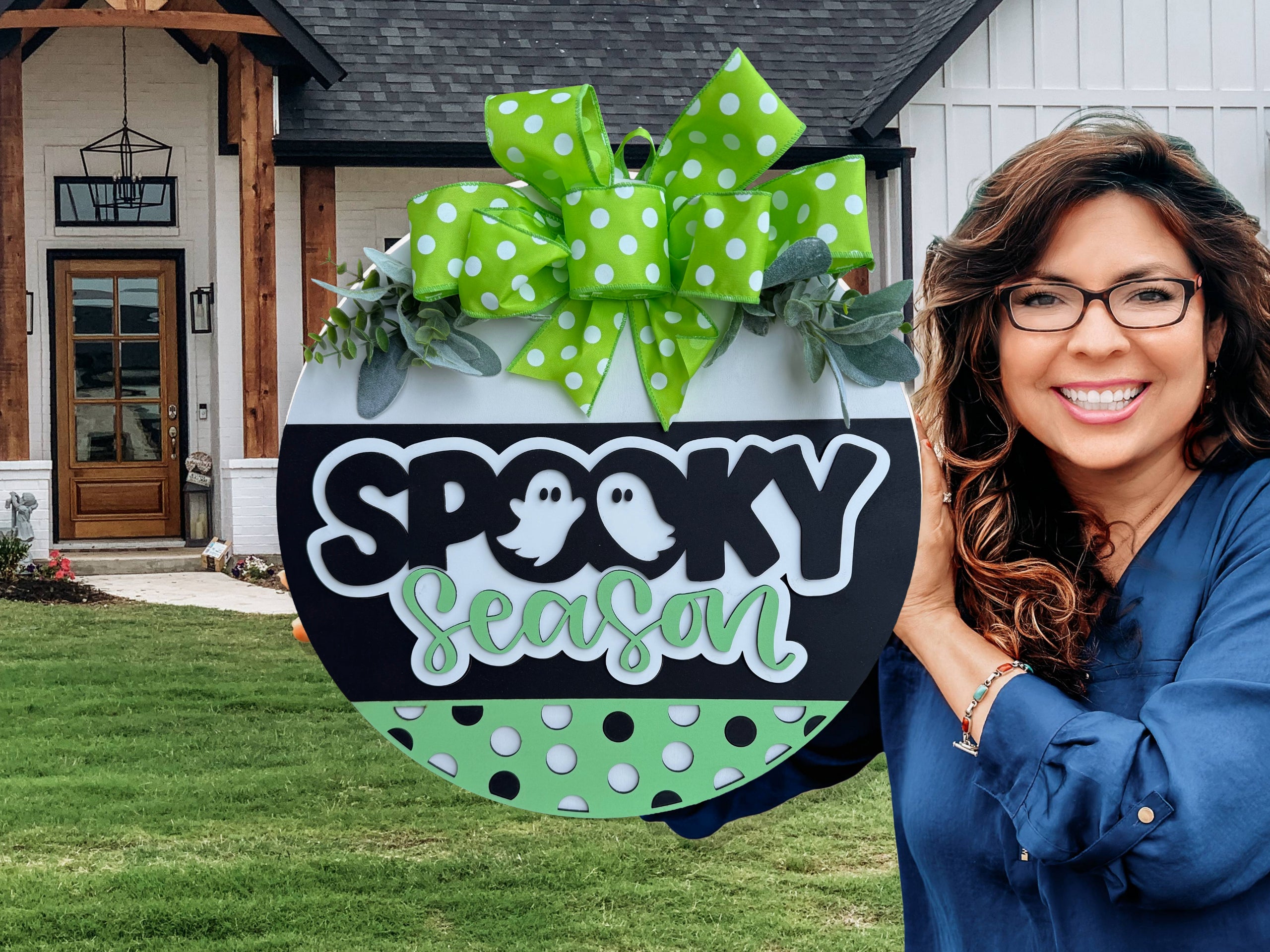 A woman with long curly hair and glasses smiles, holding a round sign decorated with green and white polka dots, leaves, and a bow. The sign reads Spooky Season with two ghost illustrations, in front of a house.