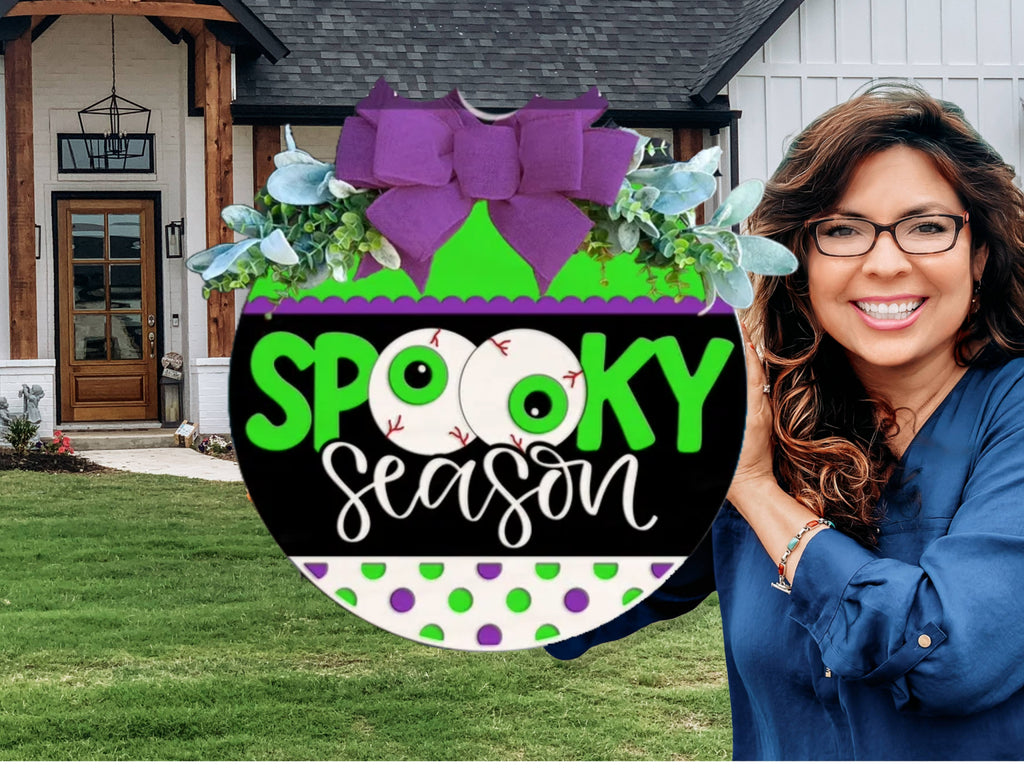 A smiling woman with long brown hair and glasses holds a round Spooky Season sign decorated with purple bows, faux greenery, and eyeballs, standing in front of a modern house with a green lawn.