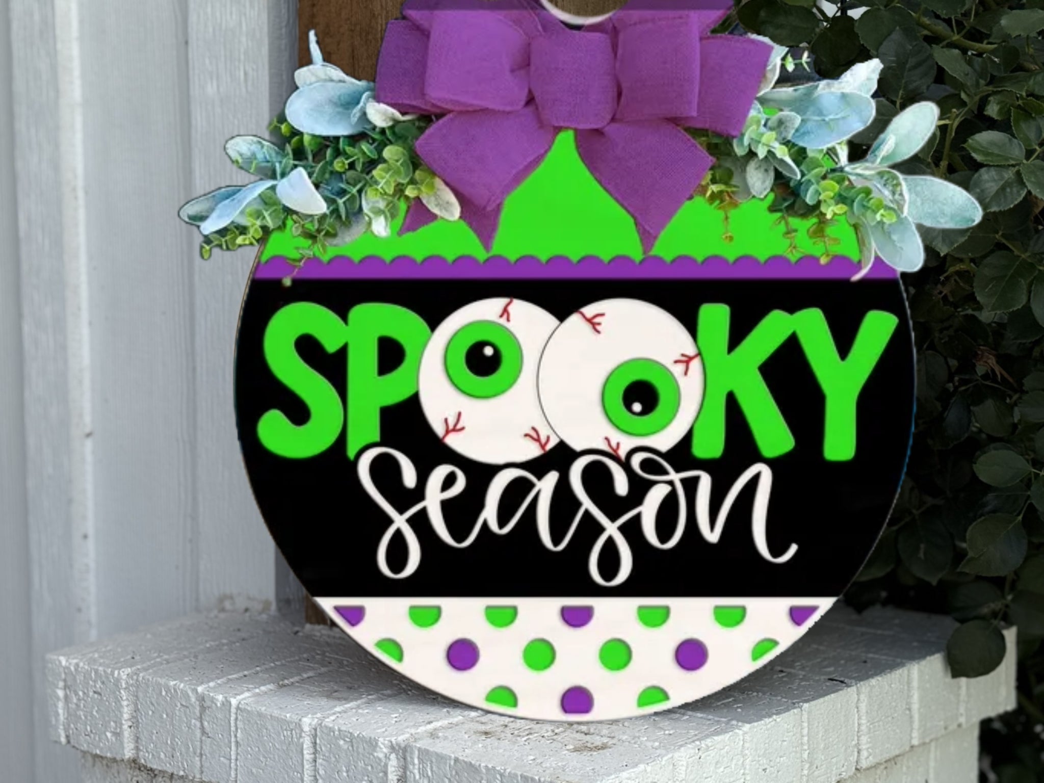 A round sign decorated with a purple bow and greenery reads Spooky Season in bright green and white letters, with two eyeballs replacing the Os in spooky and green and purple polka dots at the bottom.