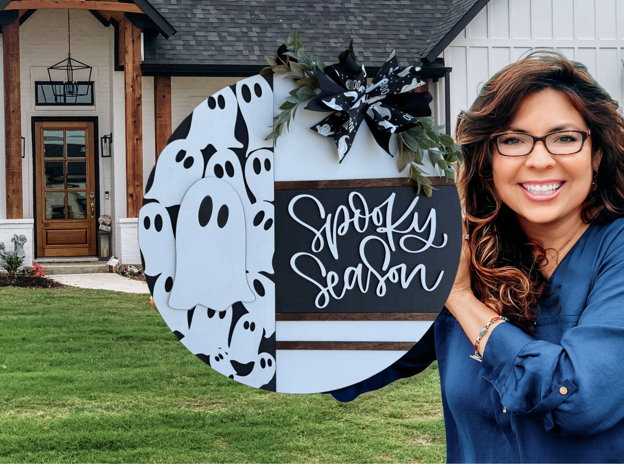 A round wooden sign with Spooky Season written on one half and a pattern of white cartoon ghosts on the other, decorated with a black and white ribbon and greenery, leaning on a white brick surface outdoors.