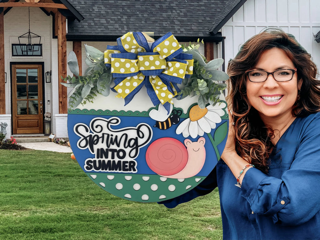 Woman smiling and holding a decorative circular sign. The sign features a design with a snail, bee, and daisy, and reads Spring Into Summer. Its adorned with a bow made of polka dot and striped ribbons. In the background is a house with a wooden entrance and lawn.