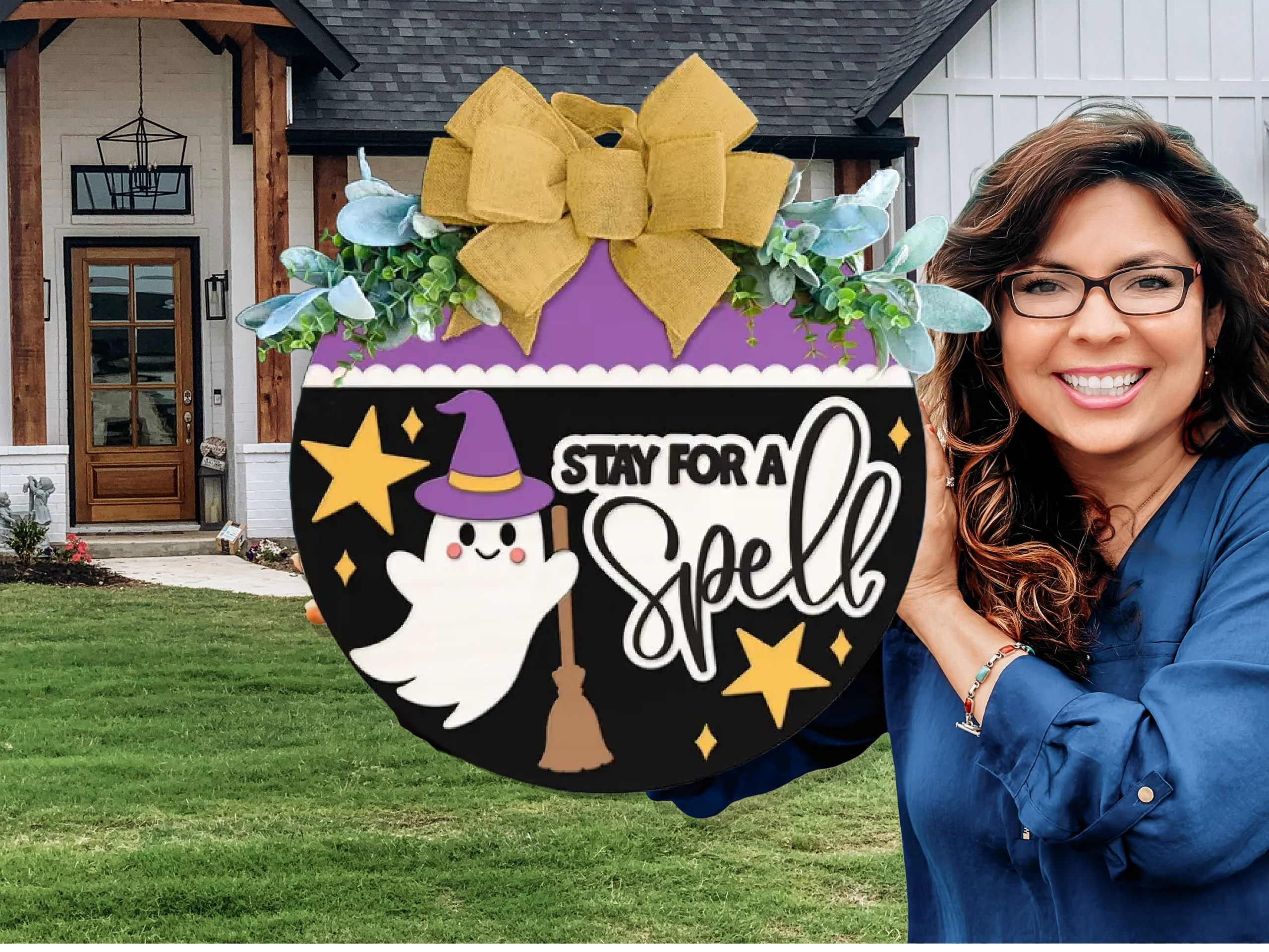 A smiling woman holds a round Halloween sign decorated with a ghost in a witch hat, stars, a broom, and the text Stay for a Spell. The sign has a yellow bow and greenery, and she stands in front of a modern house.