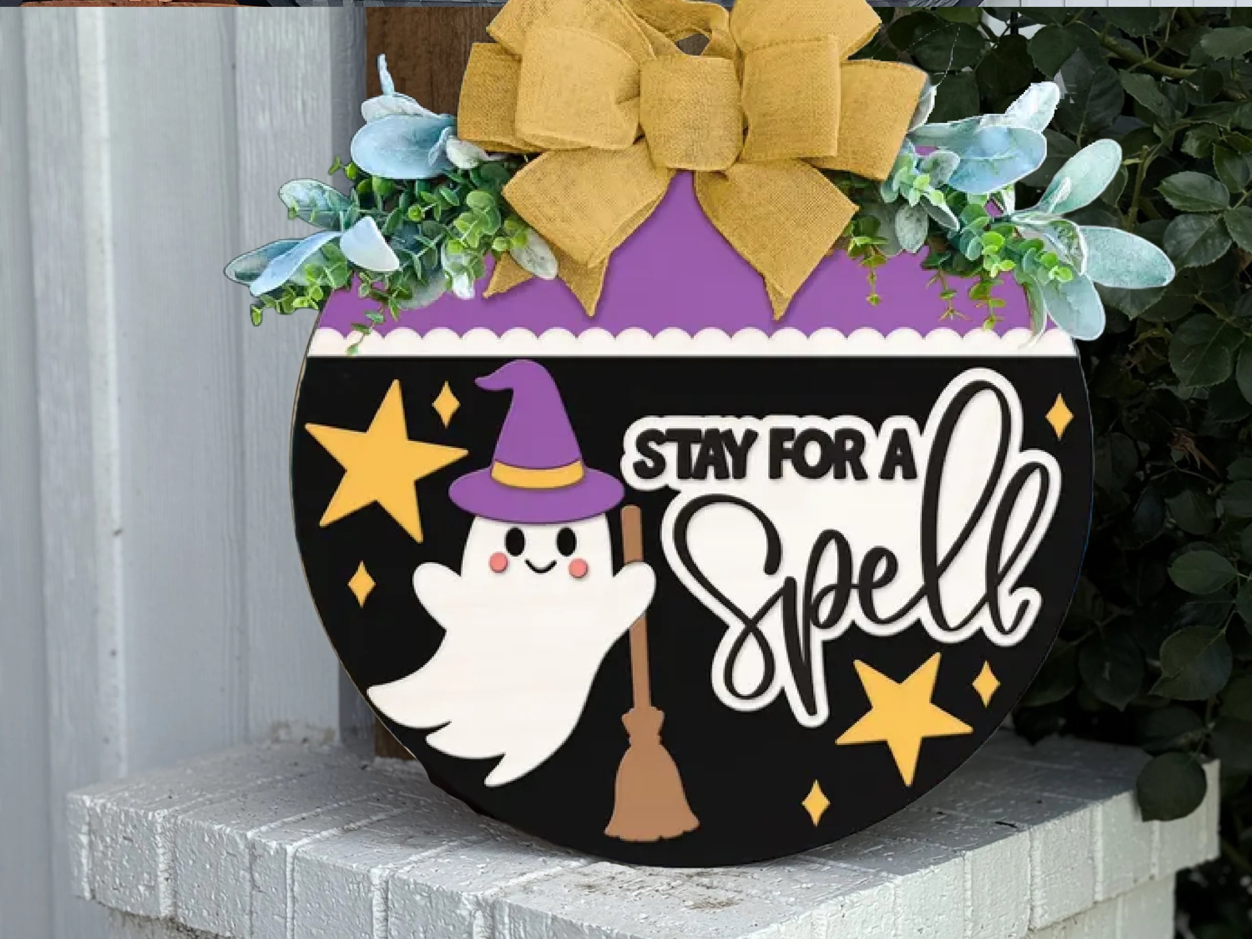 A round Halloween sign with a yellow bow, greenery, a ghost in a witch hat holding a broom, yellow stars, and the words Stay for a Spell in playful fonts, displayed on a white brick ledge.