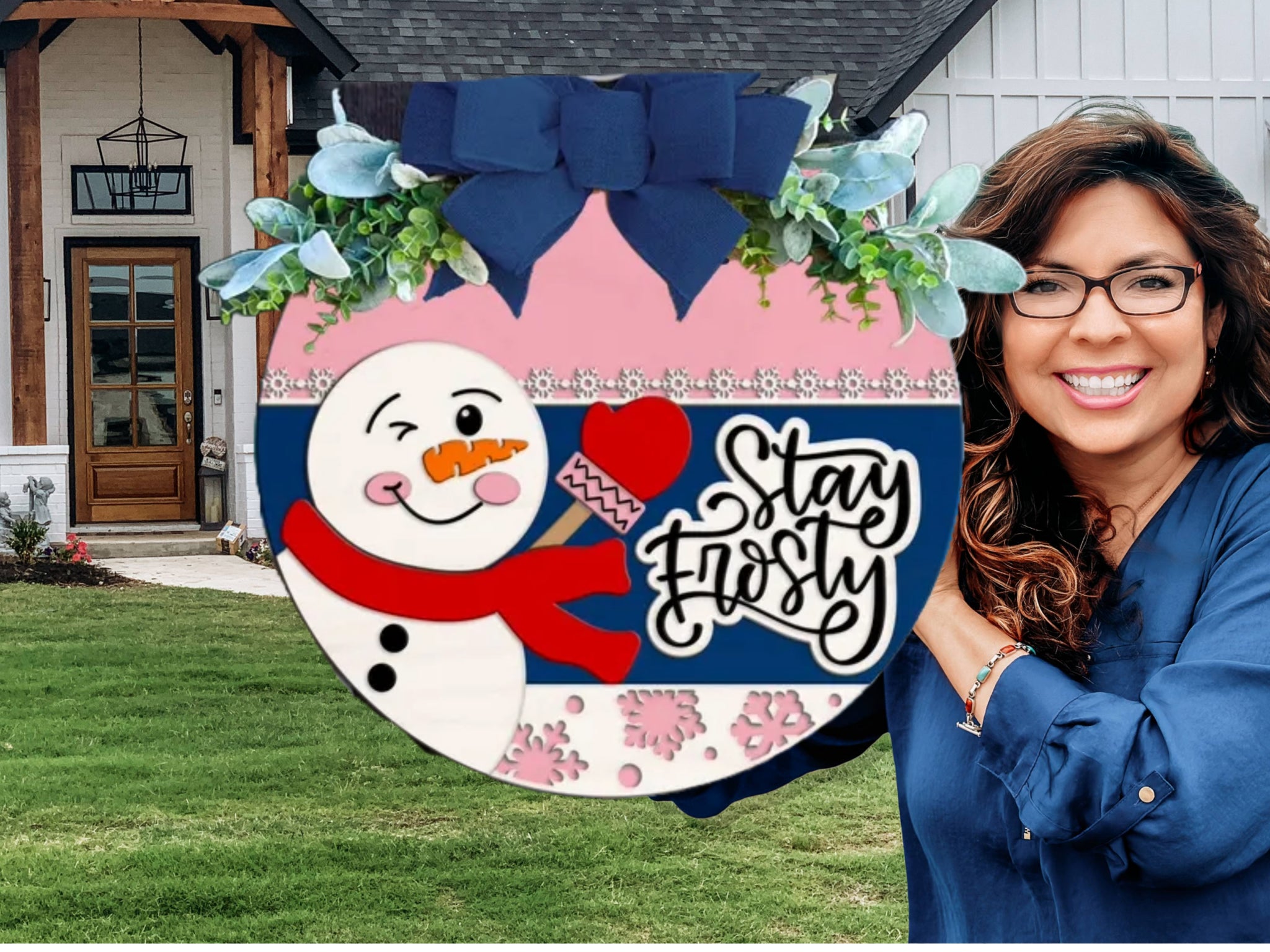 A smiling woman with glasses holds a round winter-themed sign featuring a cheerful snowman, floral decorations, and the words Stay Frosty in front of a modern house with a green lawn.