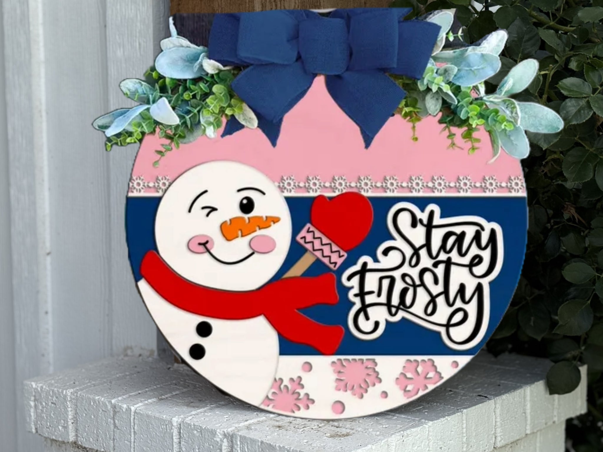 A round winter sign features a winking snowman with a red scarf and mittens, floral accents, a large blue bow, and the words “Stay Frosty” in script, displayed on a white brick surface near greenery.