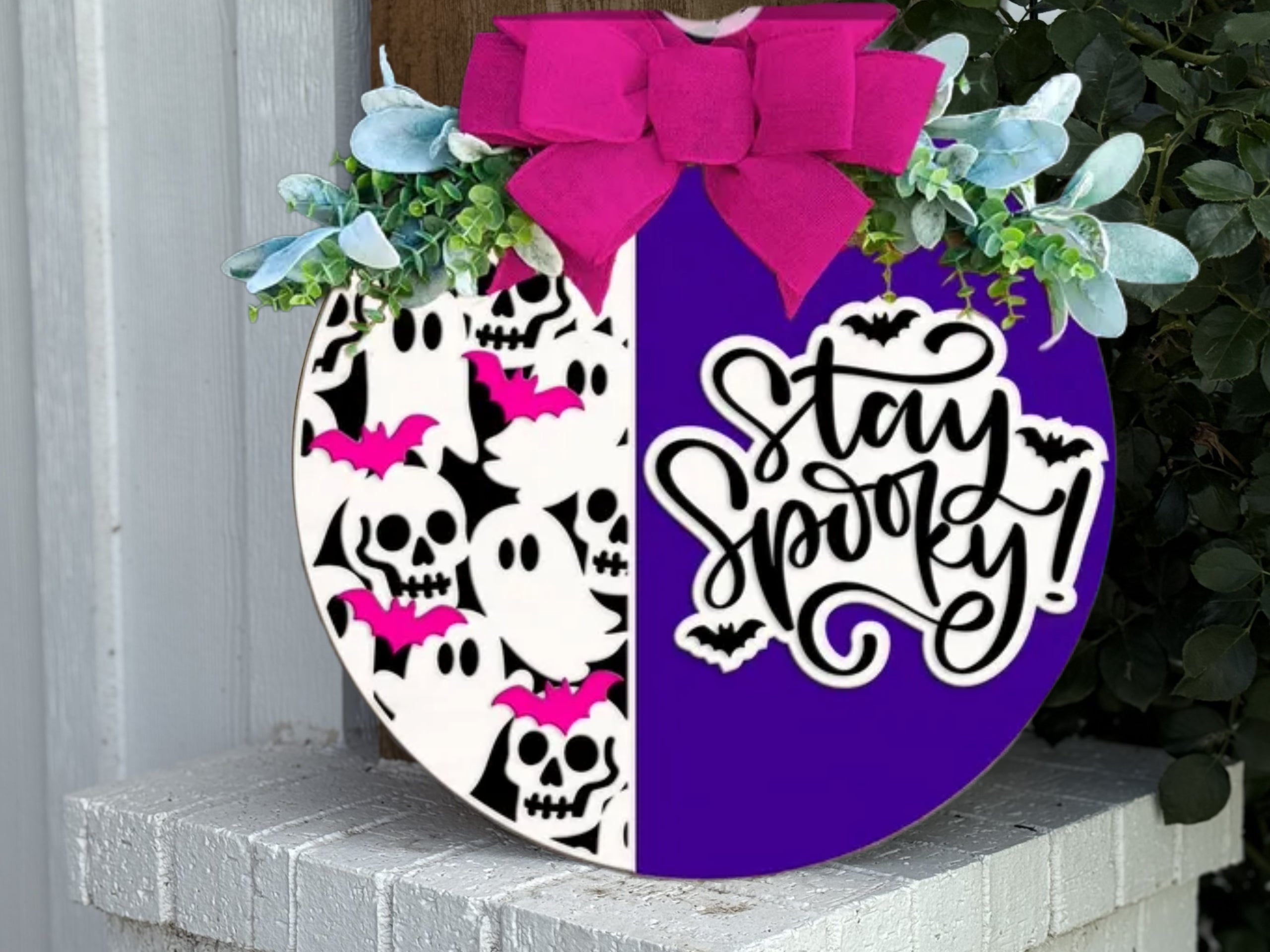 A round Halloween sign decorated with a pink bow, greenery, skulls, ghosts, pink bats, and the phrase Stay Spooky! in black script on a purple and white split background.