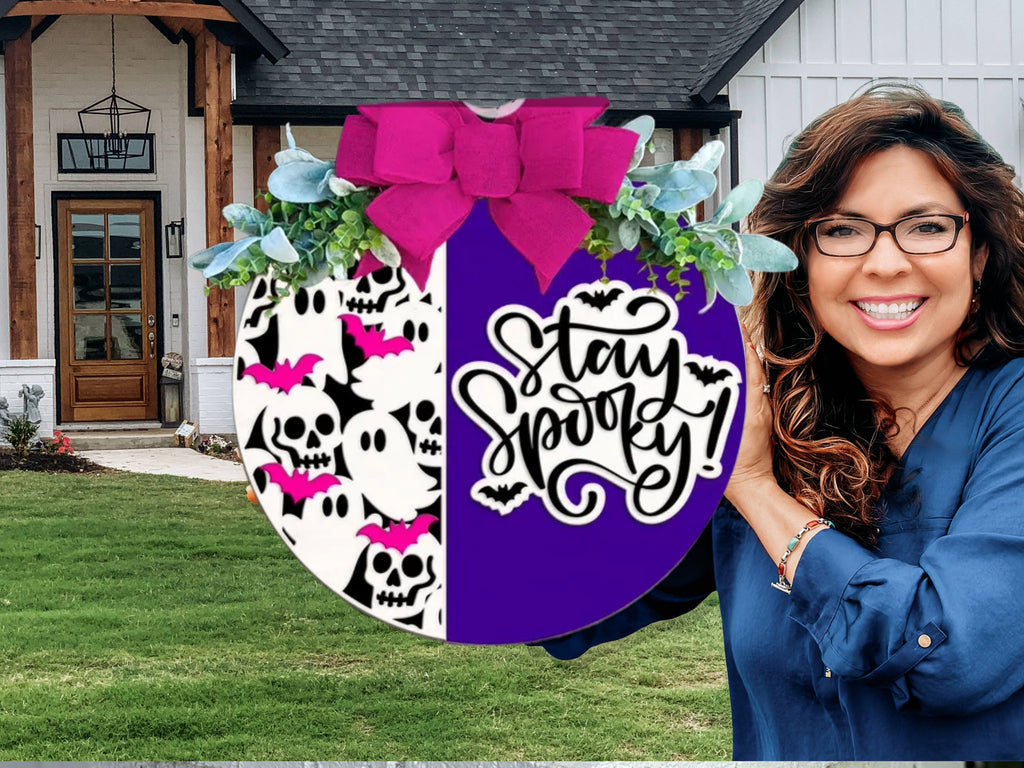 A round Halloween sign decorated with a pink bow, greenery, skulls, ghosts, pink bats, and the phrase Stay Spooky! in black script on a purple and white split background.