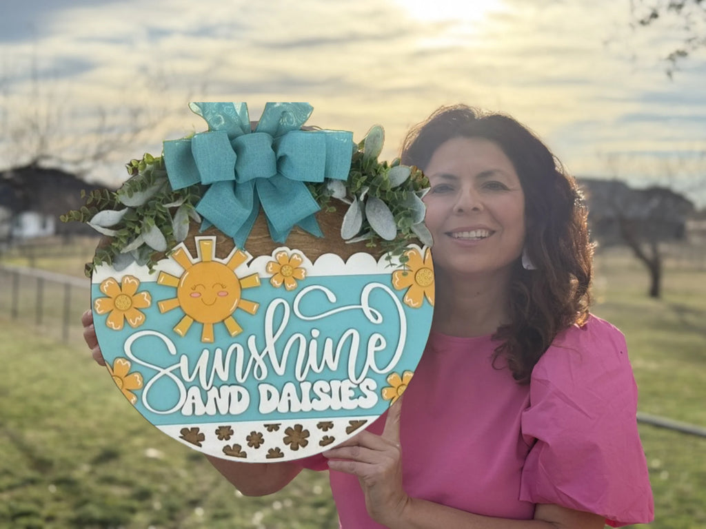 A woman in a pink shirt holds a decorative wreath featuring yellow daisies, a stylized sun, and the words Sunshine and Daisies in white script. The wreath is adorned with a large teal bow. The background shows a grassy field and a soft, cloudy sky with the sun setting.