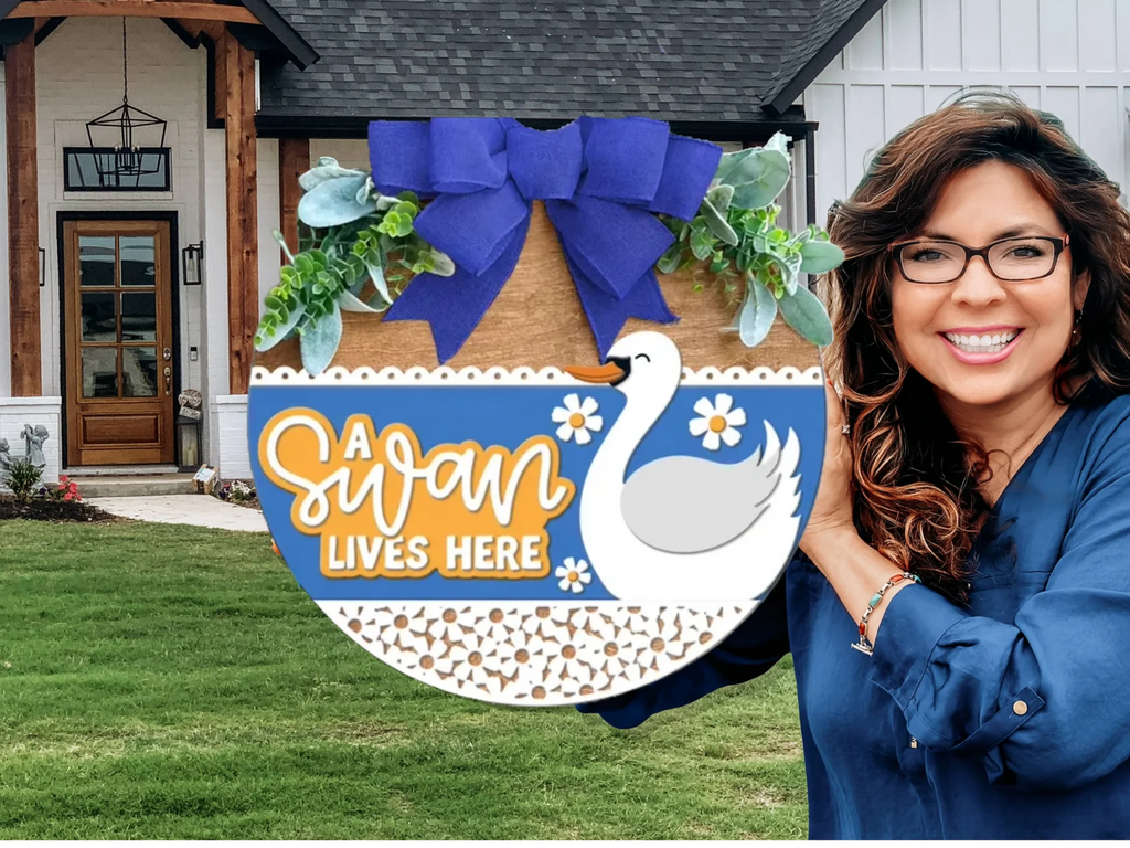 A smiling woman with long brown hair and glasses holds a decorative sign with a swan, blue bow, and greenery that reads A swan lives here, standing in front of a modern house.