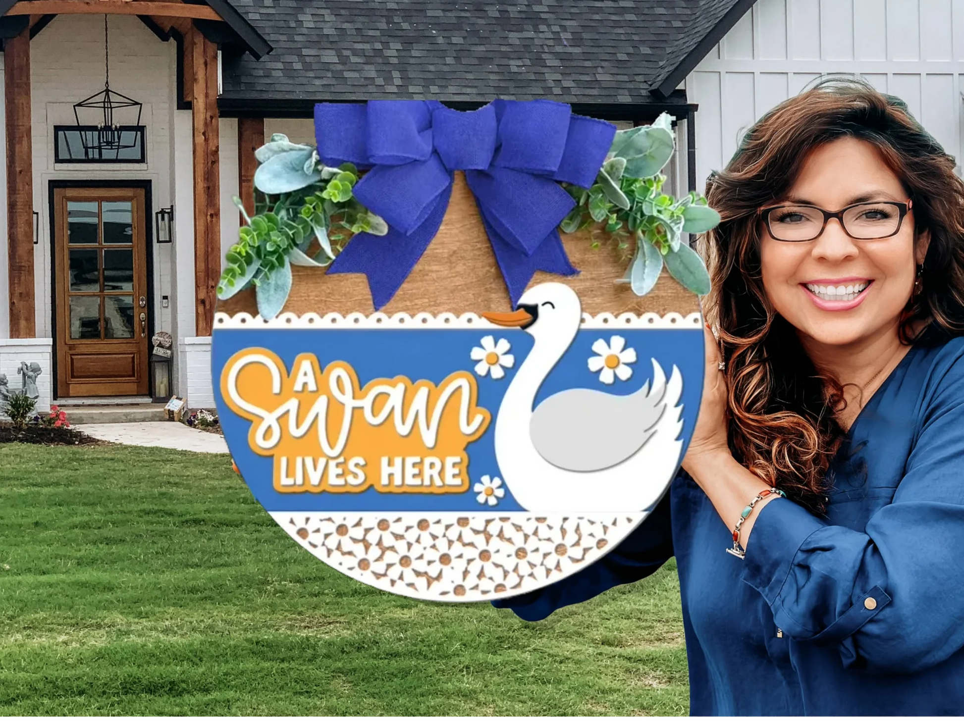 A smiling woman with long brown hair and glasses holds a decorative sign with a swan, blue bow, and greenery that reads A swan lives here, standing in front of a modern house.