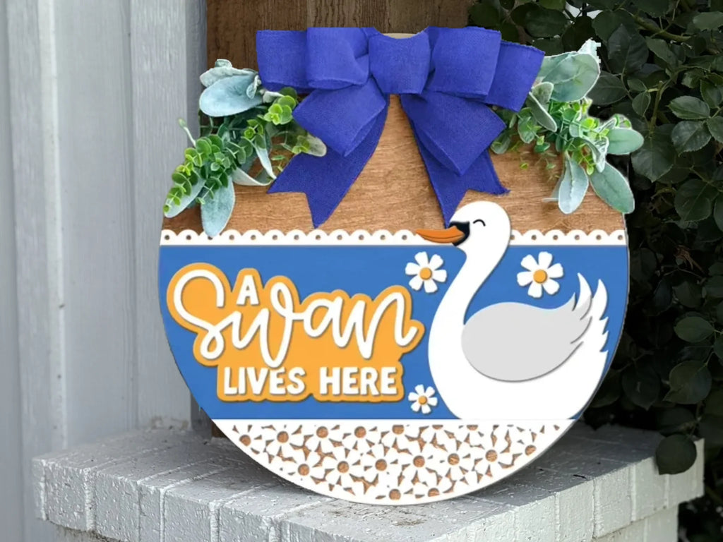A decorative round wooden sign with a blue bow, greenery, a white swan illustration, daisies, and the text “A Swan Lives Here,” placed on a white porch step.