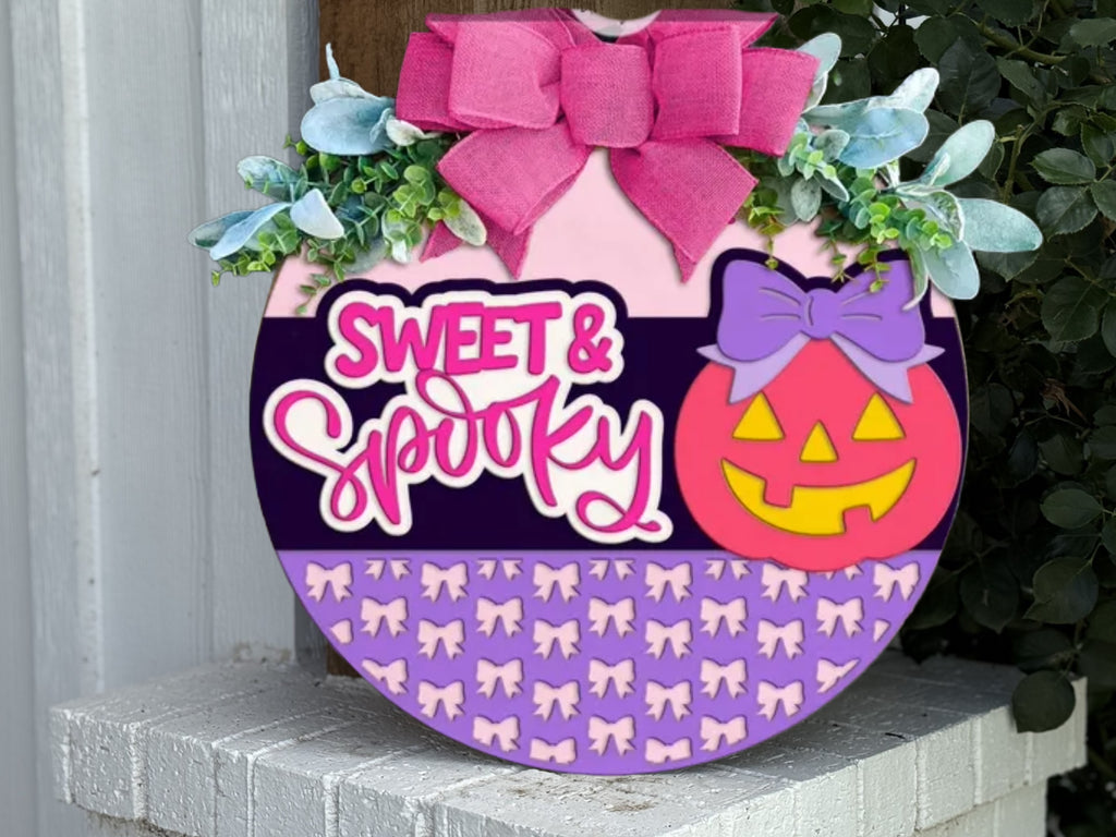 A round Halloween sign decorated with a pink bow, greenery, and a smiling jack-o’-lantern with a purple bow. The sign reads “Sweet & Spooky” and features a pattern of small purple bows along the bottom.