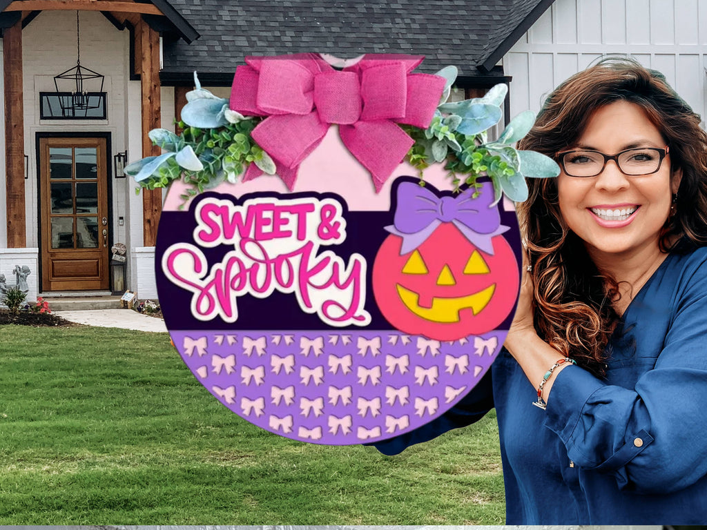 A round Halloween sign decorated with a pink bow, greenery, and a smiling jack-o’-lantern with a purple bow. The sign reads “Sweet & Spooky” and features a pattern of small purple bows along the bottom.