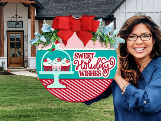 A festive round sign with a red bow, greenery, and three cupcakes under a dome. The sign reads Sweet Holiday Wishes in red letters on a teal, red, and white background with diagonal stripes.