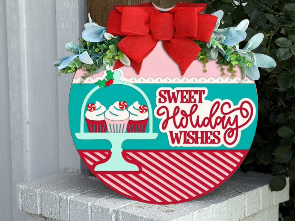 A festive round sign with a red bow, greenery, and three cupcakes under a dome. The sign reads Sweet Holiday Wishes in red letters on a teal, red, and white background with diagonal stripes.