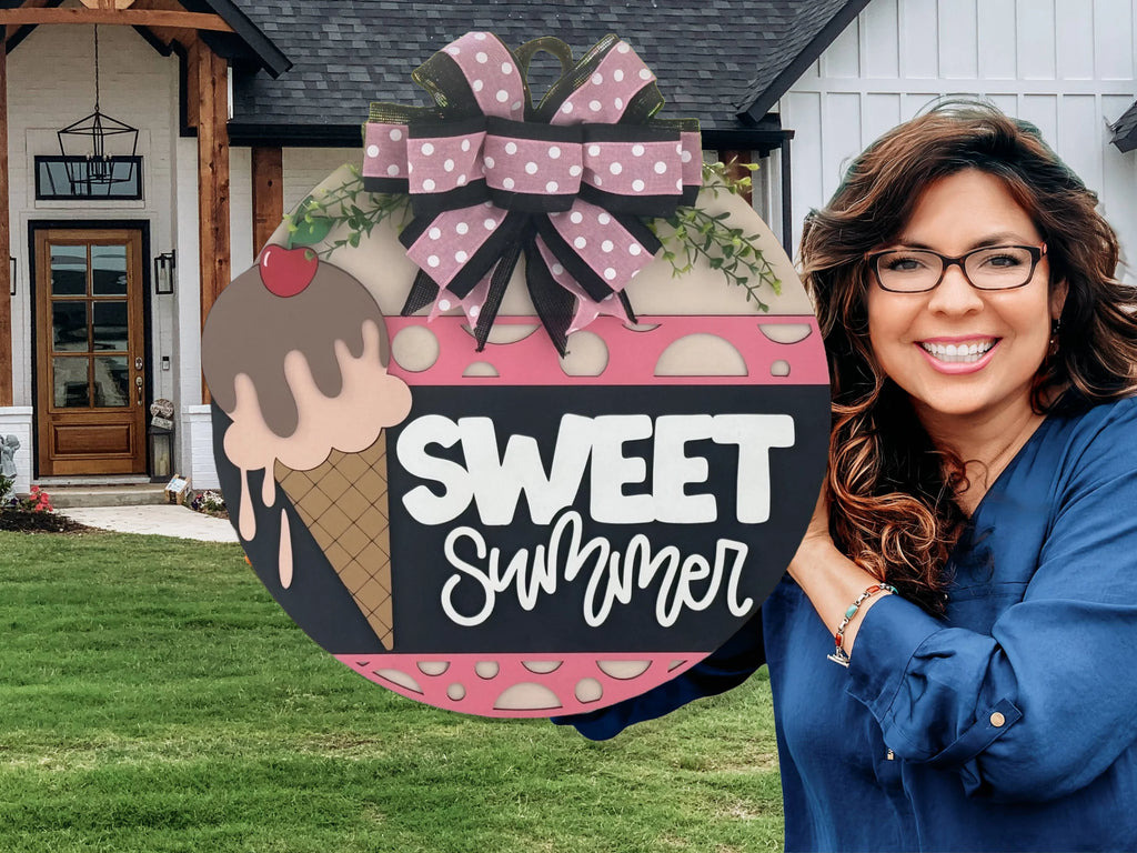 A smiling woman with glasses and wavy hair stands outside a house, holding a large round sign decorated with a pink ice cream cone, bow, and the words “SWEET SUMMER” in white letters against a pink and black background.