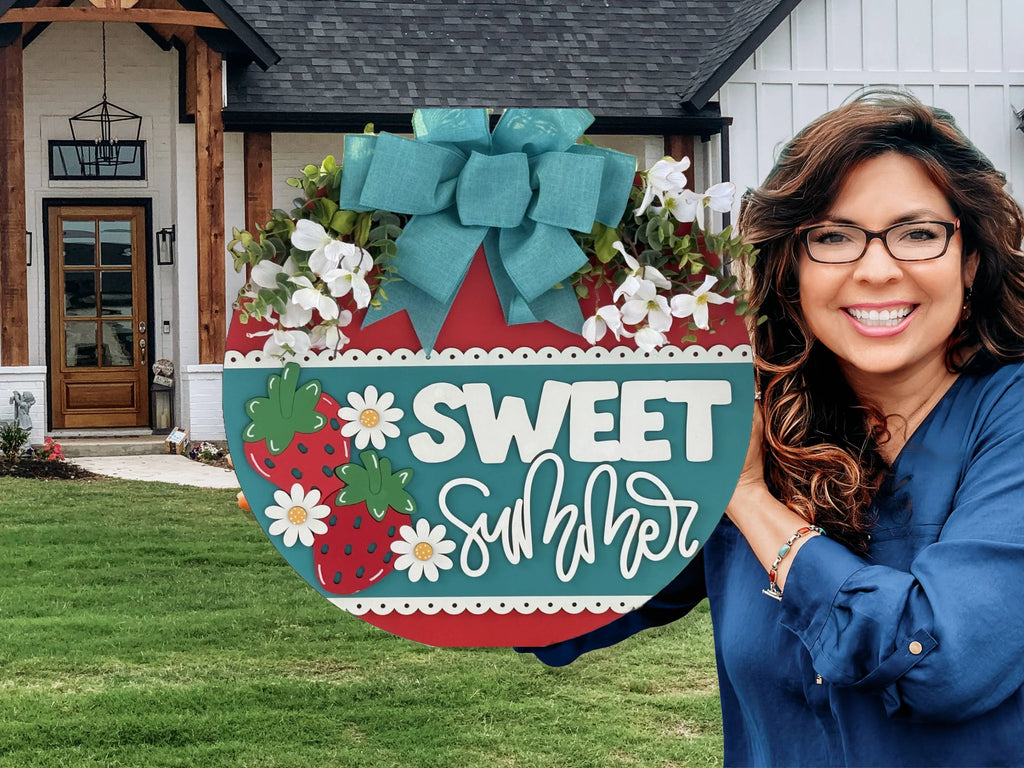 A smiling woman with long brown hair and glasses holds a round, colorful sign reading “Sweet Summer” with strawberries, daisies, and a teal bow. She stands in front of a modern house with white siding, wooden beams, and a neat green lawn.