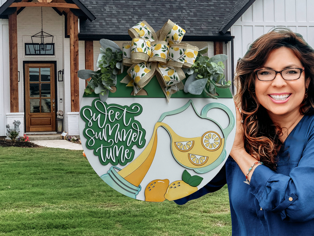 A smiling woman with wavy brown hair and glasses holds a round summer-themed sign with lemons, lemonade, and the words sweet summer time in front of a modern farmhouse.
