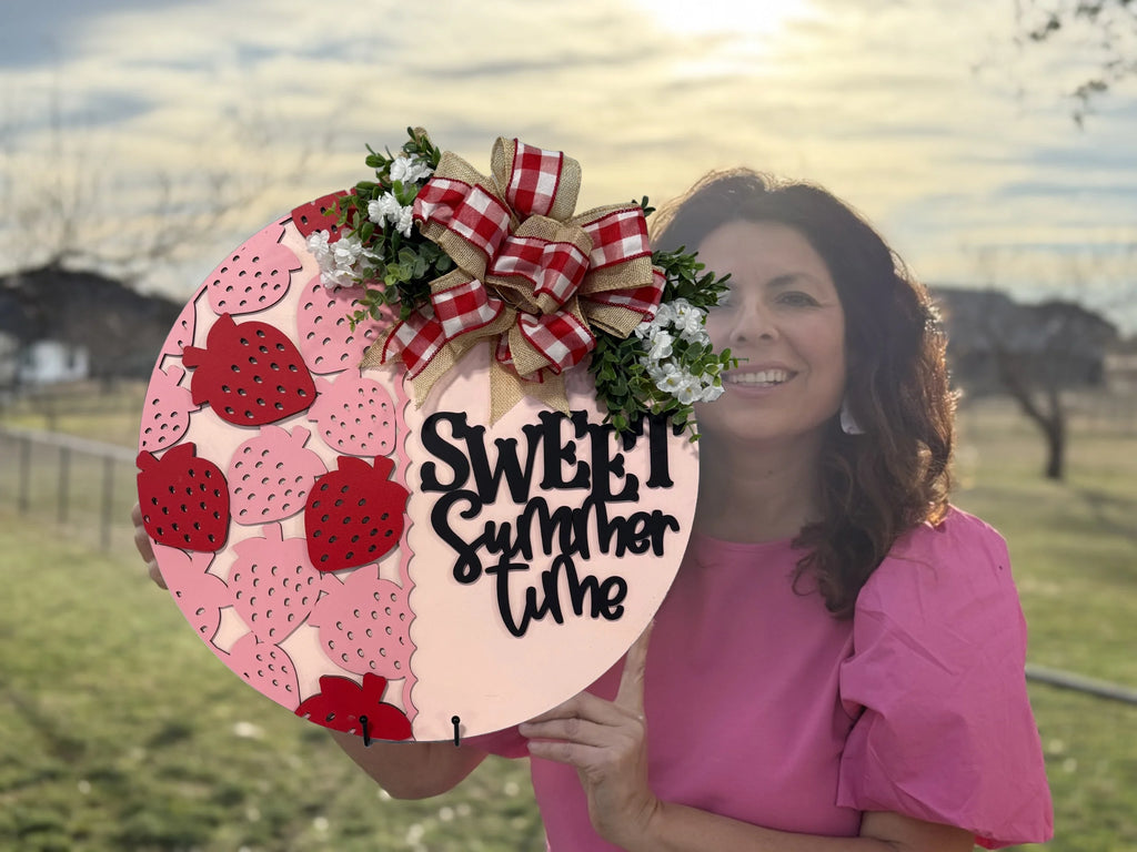 A woman in a pink blouse holds a circular sign with the text Sweet Summertime in black. The sign features a pattern of strawberries and is decorated with a red and white plaid bow and small white flowers. The background shows a grassy field and a cloudy sky.