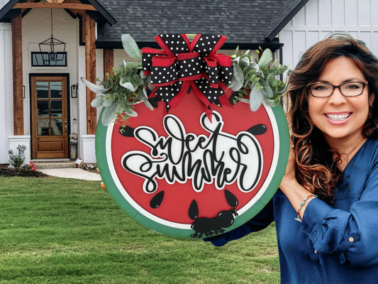A woman with long hair and glasses stands outside a modern house holding a round sign. The sign resembles a watermelon slice with the text Sweet Summer written on it, adorned with a polka dot bow and greenery. The house has a wooden front door and white siding.