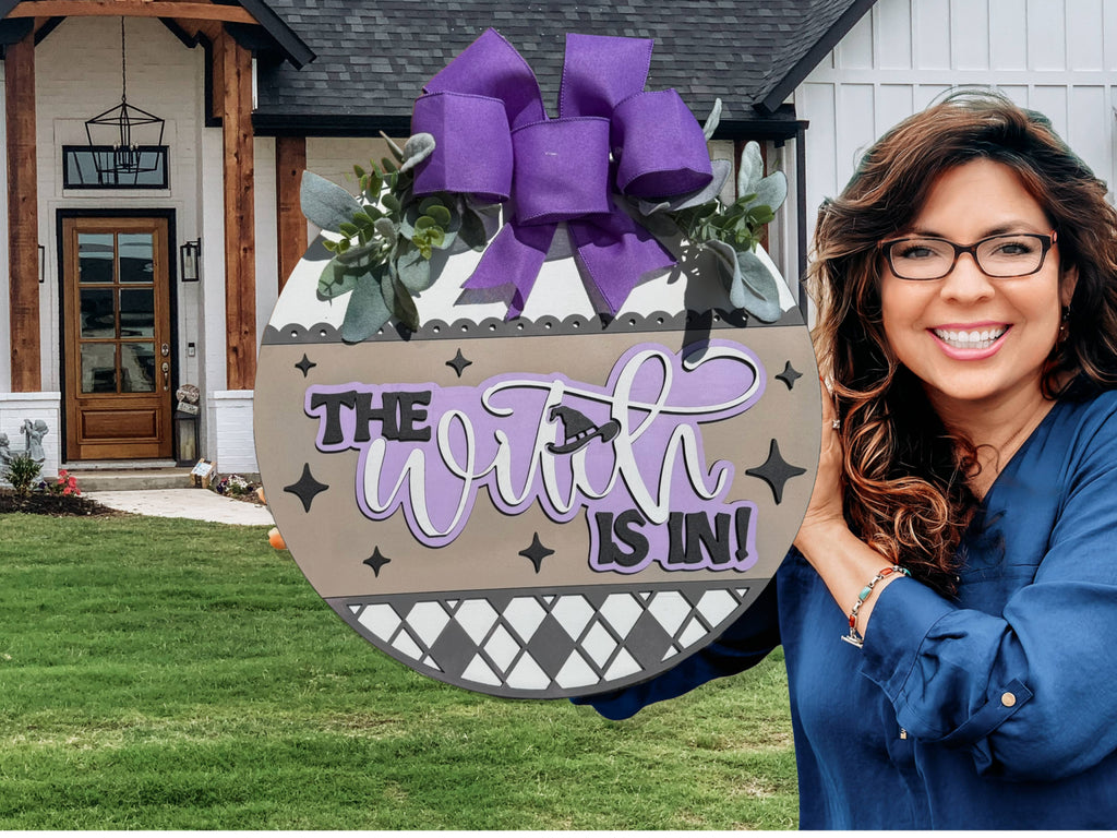 A smiling woman with long brown hair and glasses holds a round sign decorated with a purple bow and greenery. The sign reads The Witch is In! in black and purple lettering. She stands in front of a modern house.