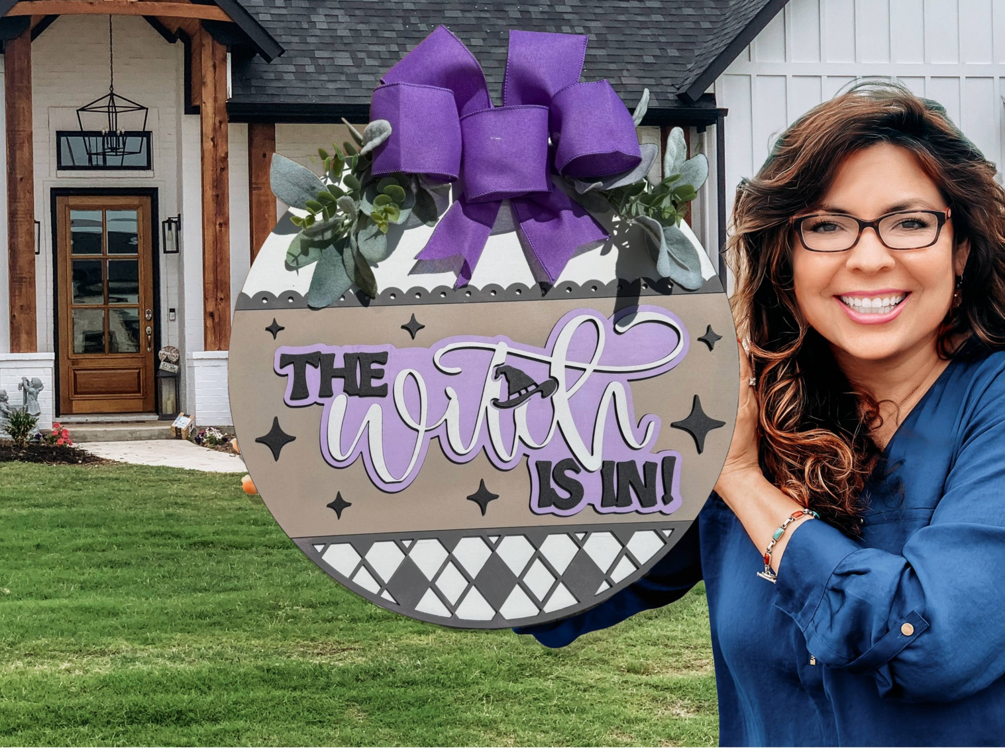 A smiling woman with long brown hair and glasses holds a round sign decorated with a purple bow and greenery. The sign reads The Witch is In! in black and purple lettering. She stands in front of a modern house.