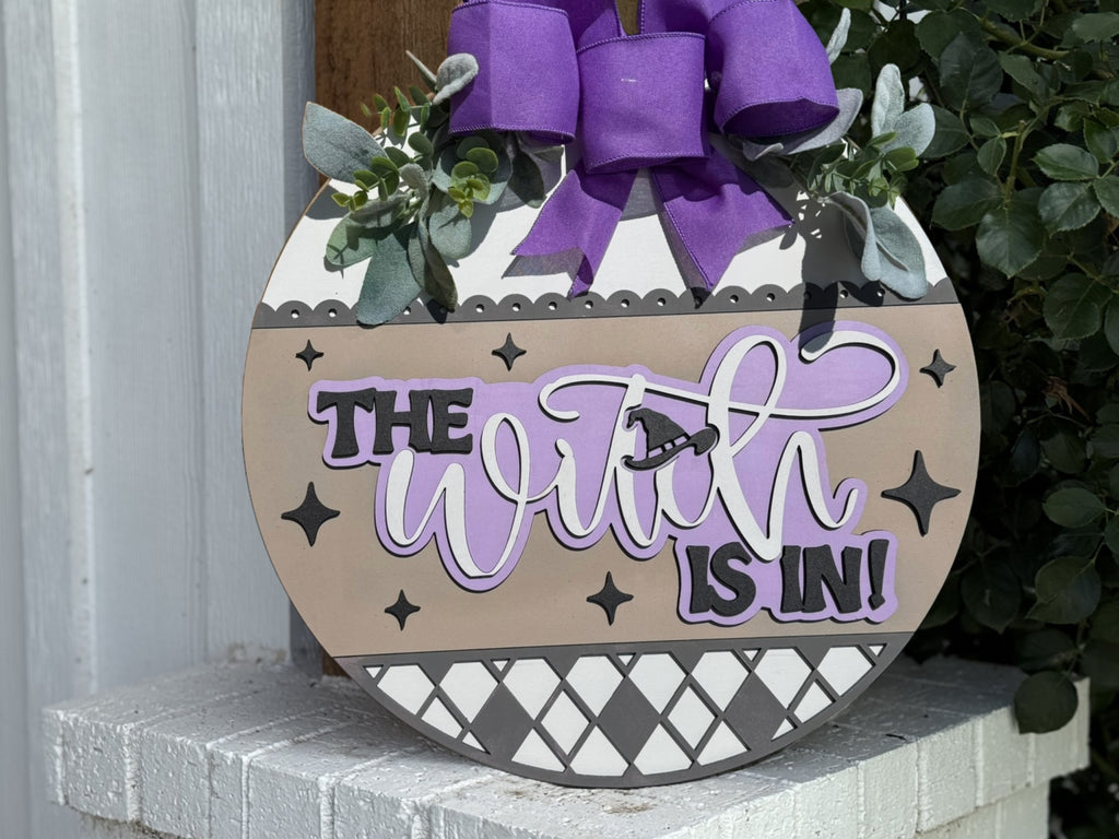 A round decorative sign with a purple bow and leaves reads The Witch is In! in bold and cursive letters, accented by star shapes and patterns, leaning against a white brick surface.