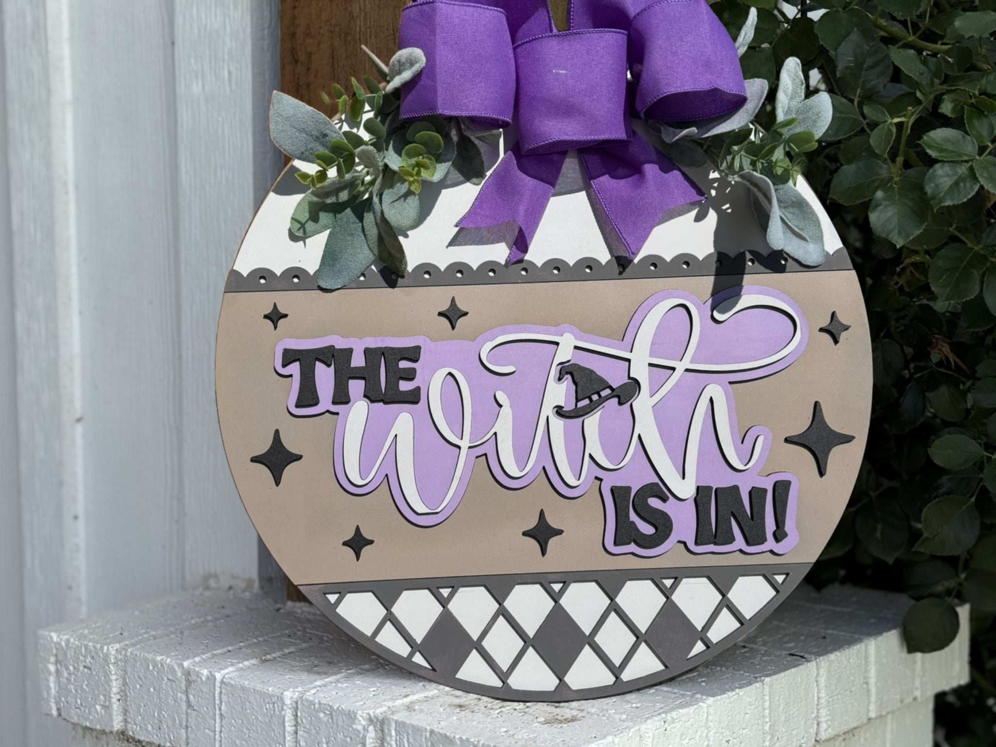 A round decorative sign with a purple bow and leaves reads The Witch is In! in bold and cursive letters, accented by star shapes and patterns, leaning against a white brick surface.