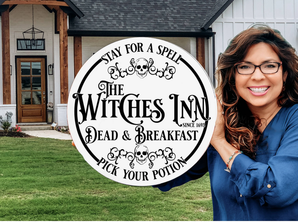 A smiling woman with wavy hair holds a round sign that reads: The Witches Inn. Dead & Breakfast. Stay for a spell. Pick your potion. Since 1693, standing in front of a modern house with wooden accents.