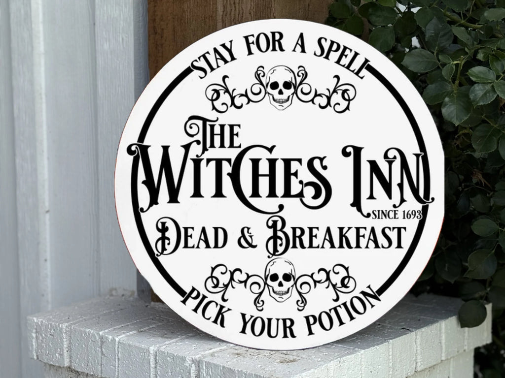 A round sign reads: “Stay for a Spell. The Witches Inn. Dead & Breakfast. Pick Your Potion. Since 1693,” with skull graphics, leaning on a white brick wall next to green leaves.