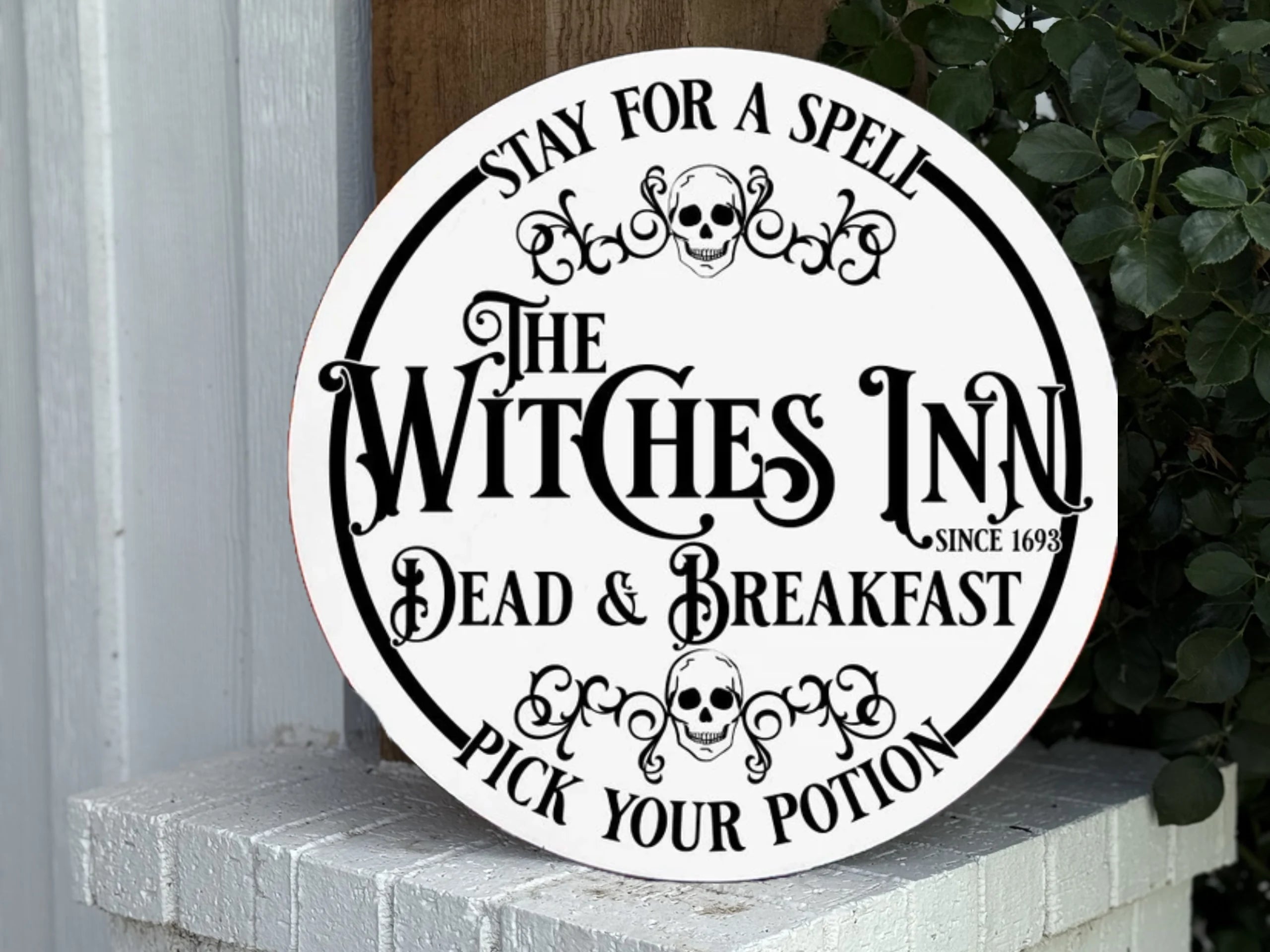 A round sign reads: “Stay for a Spell. The Witches Inn. Dead & Breakfast. Pick Your Potion. Since 1693,” with skull graphics, leaning on a white brick wall next to green leaves.