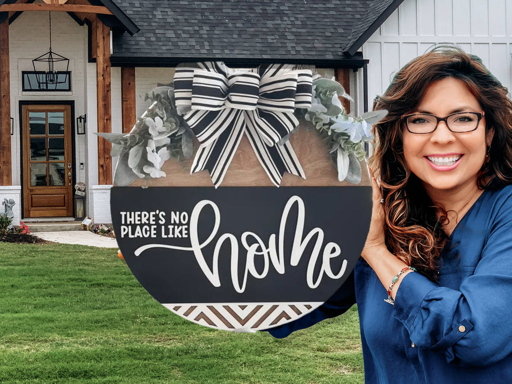 A smiling woman with long brown hair and glasses holds a round sign decorated with a striped bow and greenery that reads Theres no place like home, standing in front of a modern farmhouse-style house.