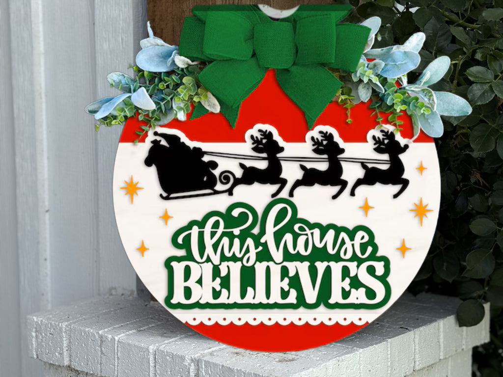 A festive sign shaped like an ornament features Santa’s sleigh and reindeer silhouettes, green foliage, and a large green bow. It reads This House Believes in white and green letters.