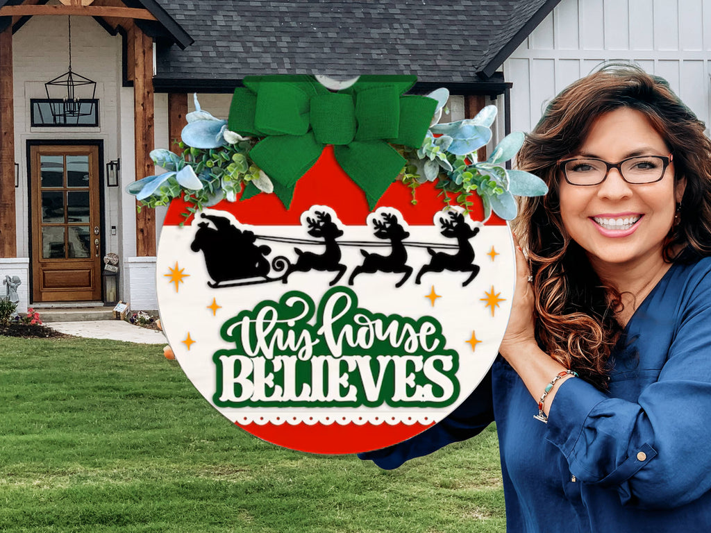 A smiling woman holds a large ornament-shaped sign with Santa’s sleigh silhouette and the words “This House Believes” in green and white, standing in front of a modern house with a lawn.