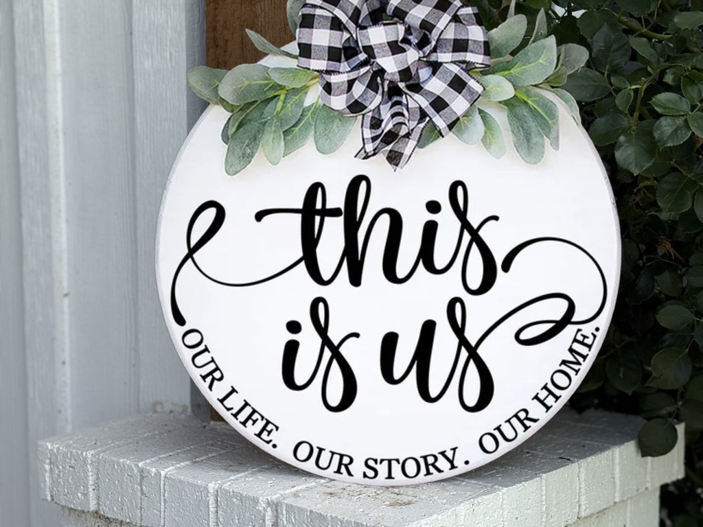 A round white sign with green leaves and a black-and-white checkered bow reads this is us in script, with OUR LIFE. OUR STORY. OUR HOME. along the bottom edge, resting on a white brick surface.