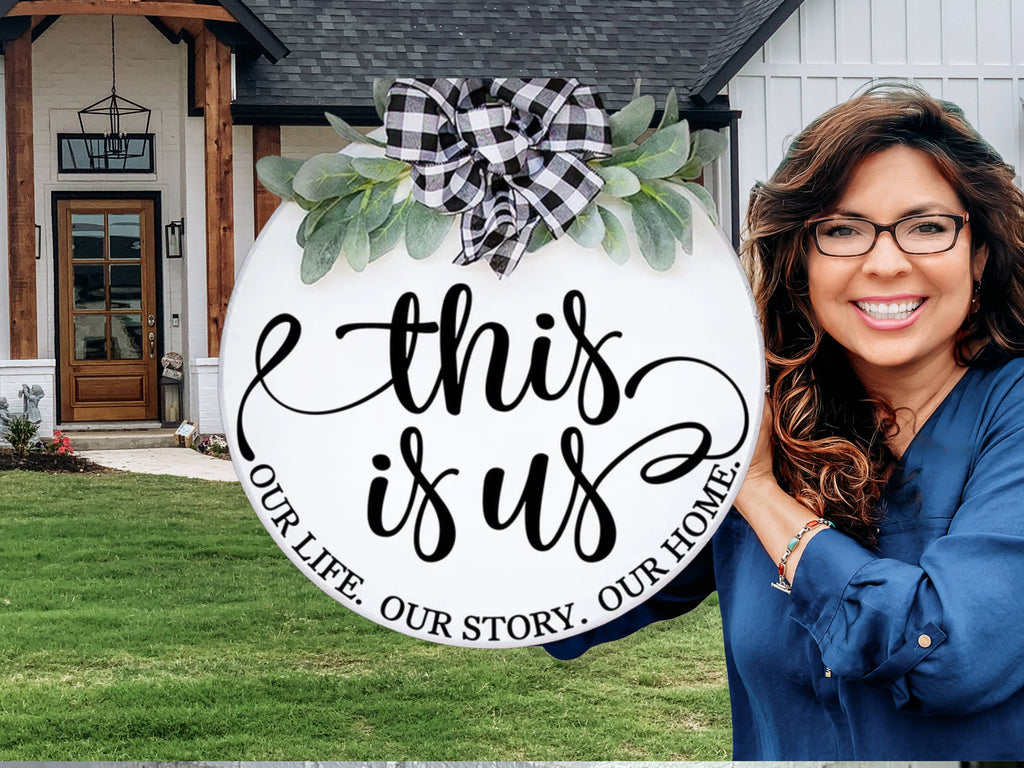 A smiling woman in glasses holds a round sign decorated with a black and white plaid bow and greenery. The sign reads, “This is us. Our life. Our story. Our home.” She stands in front of a modern house with a lawn.