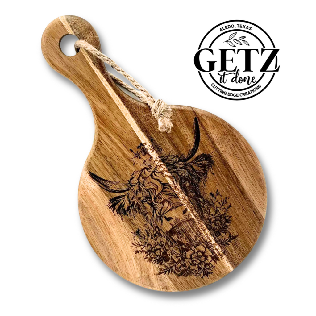 Round wooden cutting board with a carved design of a cows head, sunflowers, and foliage. A rope is looped through the handle. The text GETZ it done and Aledo, Texas, Cutting Edge Creations is printed in black on the upper right.