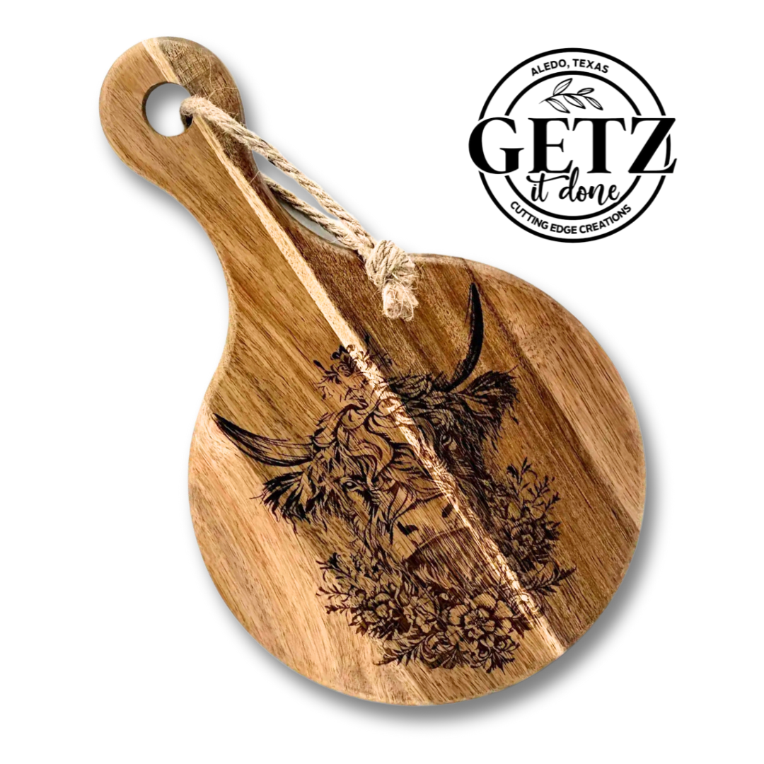 Round wooden cutting board with a carved design of a cows head, sunflowers, and foliage. A rope is looped through the handle. The text GETZ it done and Aledo, Texas, Cutting Edge Creations is printed in black on the upper right.