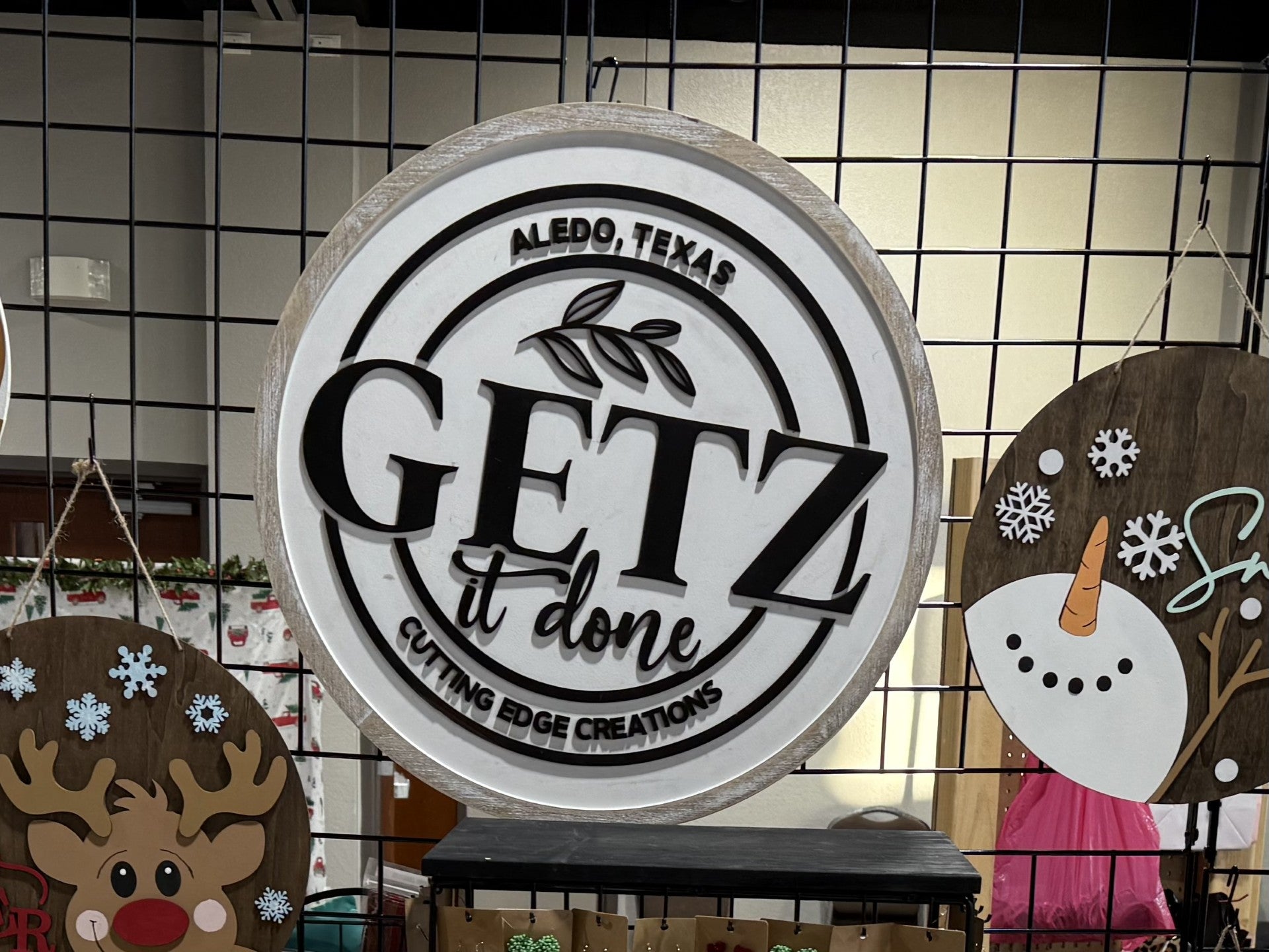 A circular sign hanging on a grid display reads GETZ it done in large black letters, with ALEDO, TEXAS and CUTTING EDGE CREATIONS in smaller text. Decorative art featuring a reindeer and a snowman is visible nearby.