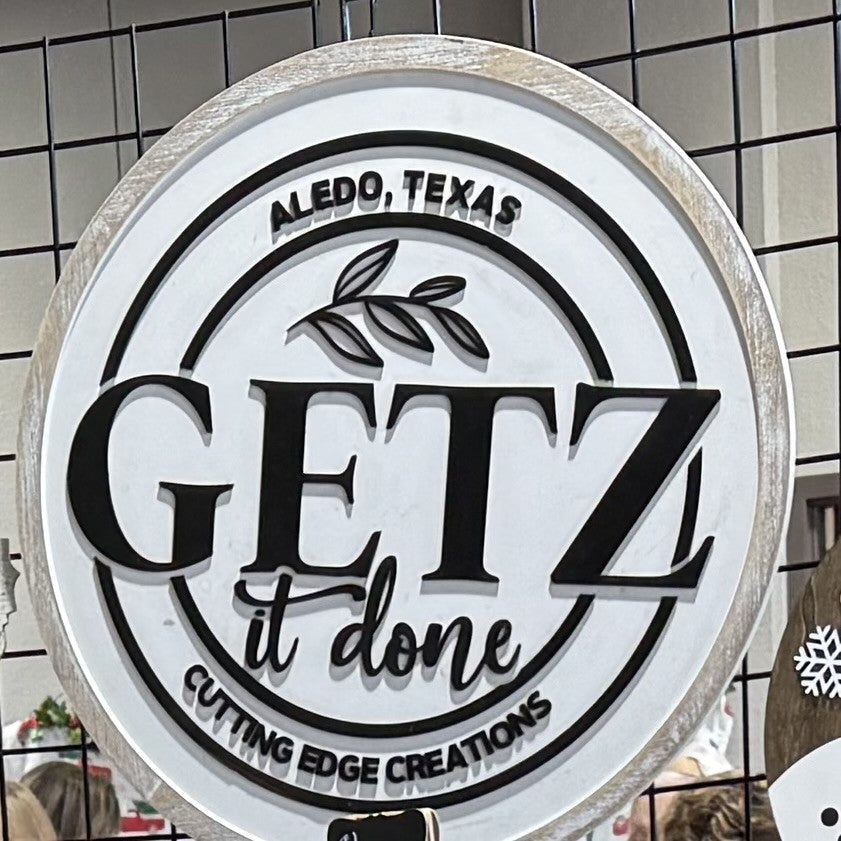 A round wooden sign with the text GETZ it done in bold black letters. The top reads Aledo, Texas and the bottom says Cutting Edge Creations. A small branch with leaves decorates the center. The sign hangs on a black grid background.
