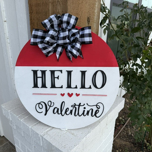 A round sign hangs on a wall featuring a red and white design with HELLO Valentine in bold black letters. Three small red hearts accent the text. Above, a black and white plaid bow decorates the top. The sign rests on a white brick column with greenery in the background.